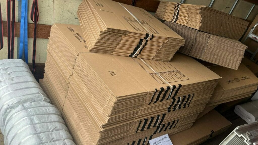 Stacks of flat cardboard moving boxes, a wrapped bundle, and a shipping label are arranged inside the back of a moving truck. Straps are hanging on the wall.