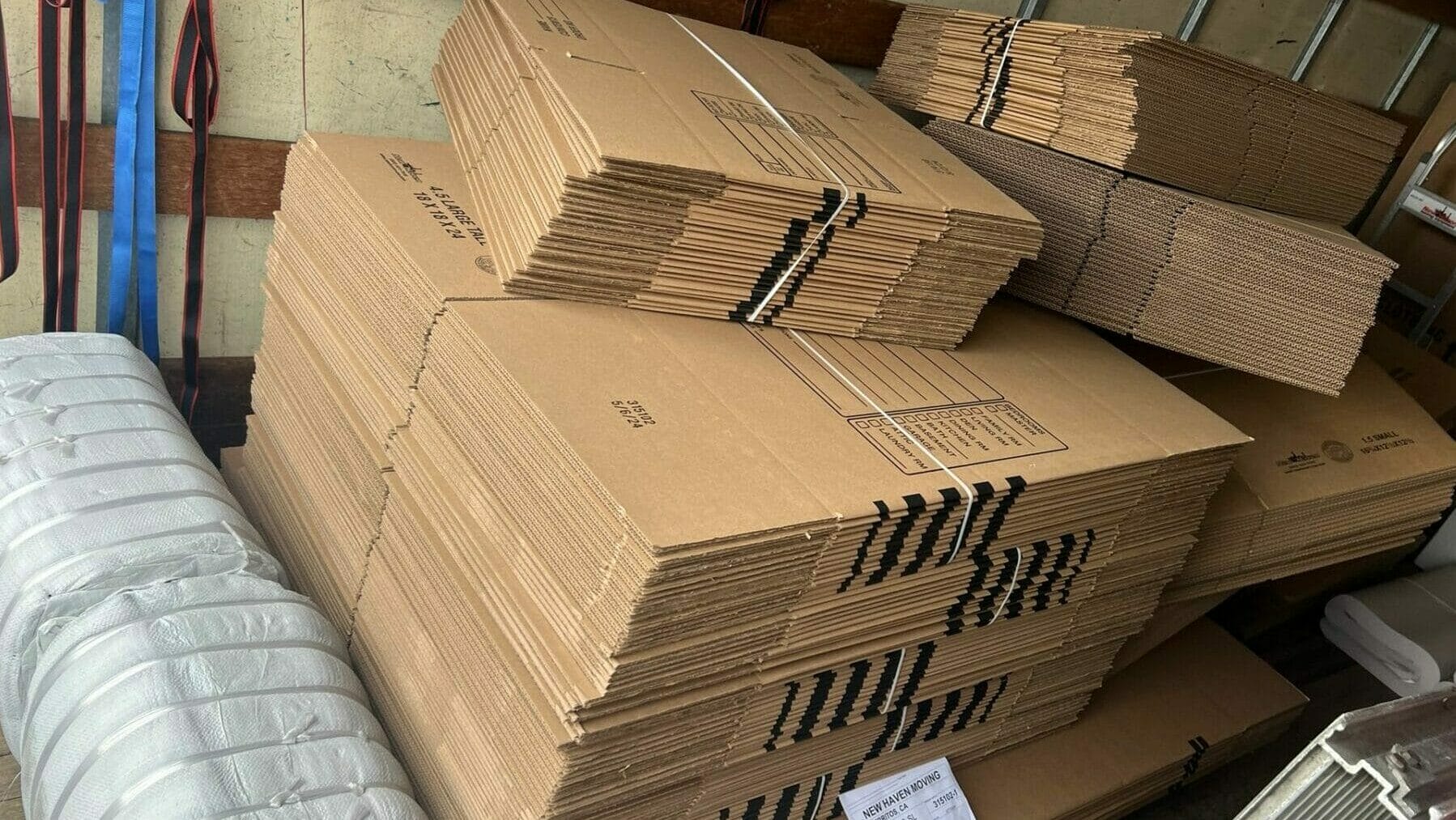 Stacks of flat cardboard moving boxes, a wrapped bundle, and a shipping label are arranged inside the back of a moving truck. Straps are hanging on the wall.