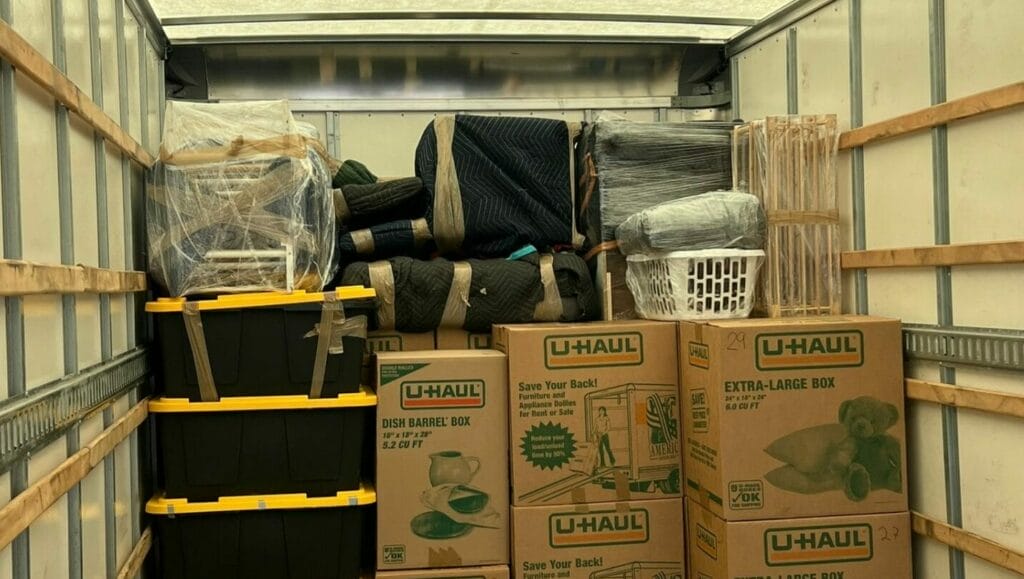 A moving truck with a neatly packed stack of U-Haul boxes, black and yellow storage bins, wrapped furniture, and rolled-up rugs against the front wall, leaving open space inside the truck.