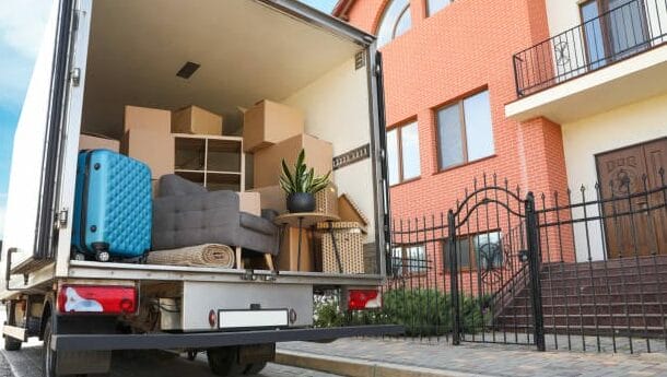 A moving truck parked outside a house with its back open, showing packed boxes, a rolled-up rug, a suitcase, and furniture being loaded or unloaded. The house has a brick exterior and a black gate.