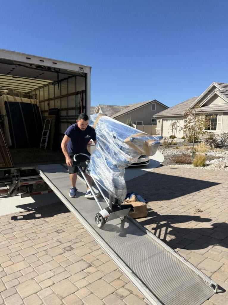 A man from a moving company pushes a dolly with a large, shrink-wrapped item down a moving truck ramp onto a driveway in a suburban neighborhood under a clear blue sky.