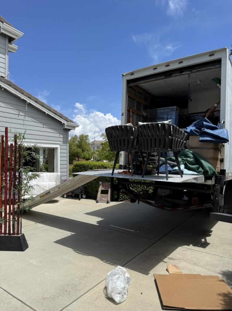 A moving truck from a professional moving company is parked in a driveway with its ramp down. Black chairs, furniture, and boxes are neatly arranged inside, showcasing the organized touch that comes with expert moving service. A house and a red bamboo plant stand are visible nearby. The sky is blue with scattered clouds.